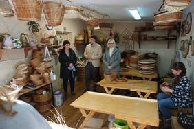 Pokaż zdjęcie: Pani zaplata wianek. Oprowadzający wraz z panem dyrektorem oglądają koszyki z wikliny, które zawieszone są przy suficie na drążku. Część koszyków bez rączek złożona jest na stole