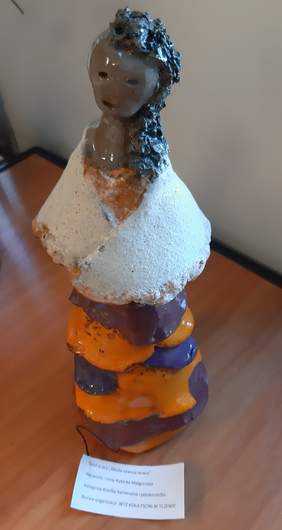 Pokaż zdjęcie: Rzeźba kobiety w sukni z falbanami w kolorach fioletu i oranżu (pomarańczy). Kobieta ma na sobie białą narzutę oraz grafitowe włosy do ramion, ułożone na lewy bok.