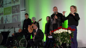 Pokaż zdjęcie: Grupa osób stoi na scenie. Po prawej stronie stoi kobieta w czarnej bluzce i granatowych spodniach, tłumacz języka migowego. Przed kobietą stoi wazon z białymi i czerwonymi różami. Po lewej stronie duży ekran z kolorową grafiką.