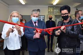 Pokaż zdjęcie: Fragment pomieszczenia, na pierwszym planie stoją dwie kobiety i w środku mężczyzna. Każdy ma w ręku nożyczki i przecina czerwona wstęgę. 