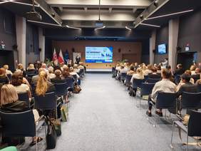Szeroki kadr nowoczesnej, symetrycznej sali konferencyjnej wypełnionej publicznością siedzącą na ciemnoniebieskich krzesłach. Rzędy krzeseł są rozdzielone szerokim, szarym przejściem pośrodku. Na końcu sali, na podwyższeniu, stoi mężczyzna przemawiający do mikrofonu. Po lewej stronie sceny widoczne są trzy flagi na stojakach (w tym flaga Polski i Unii Europejskiej). W tle znajduje się duży ekran z prezentacją konferencyjną zatytułowaną „SIEĆ WSPÓŁPRACY”. Pod sufitem wiszą profesjonalne projektory i oświetlenie.