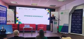 Pokaż zdjęcie: Przy mównicy z logotypem ZUS przemawia Urszula Kierznowska z Oddziału Podlaskiego PFRON. W tle znajduje się duży ekran z napisem „OTWARCIE KONFERENCJI” oraz banery instytucji, w tym ZUS, PFRON i Ministerstwa Funduszy i Polityki Regionalnej. Na scenie ustawione są czerwone fotele i stolik z butelkami wody. Po lewej stronie ekranu widoczny jest monitor z tłumaczem języka migowego. 