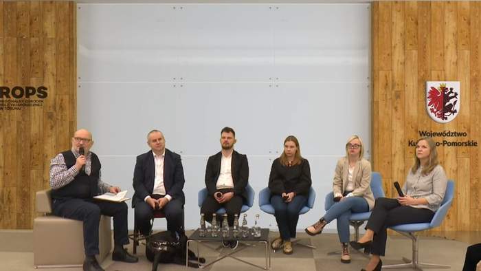 Pokaż zdjęcie: Na środku sali siedzi 6 osób, prelegencci , prowadzacy IV panel Forum pn. "Zatrudnienie osoby z niepełnosprawnością intelektualną", w tle widać logo województwa kujawsko-pomorskiego oraz ROPS-u