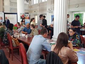 Duża sala, na niej targi pracy, część osób siedzi za stołami, pozostali chodzą, 