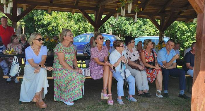 Pokaż zdjęcie: Pod drewnianym zadaszeniem na ławkach siedzą ludzie ubrani wakacyjnie, na kolorowo. W tle drzewa i zaparkowane samochody. Świeci mocne słońce.