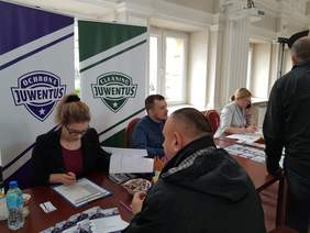 przy stole siedzi kobieta i mężczyzna, naprzeciwko nich mężczyzna, na prawo kobieta rozmawia przez telefon, przed nią widać fragment sylwetki mężczyzny