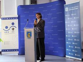 Pokaż zdjęcie: Kobieta przemawia przy mównicy z herbem Powiatu Puckiego, trzyma mikrofon. W tle niebieska ścianka z napisem „Powiat Pucki” oraz roll-upy dotyczące Roku Wsparcia Osób z Niepełnosprawnościami w Powiecie Puckim 2026.