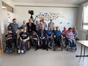 Pokaż zdjęcie: Grupa uśmiechniętych osób pozuje do zdjęcia. Są wśród nich osoby siedzące na wózkach inwalidzkich. Po środku siedzi mężczyzna w ciemnym garniturze z psem asystującym