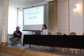 Pokaż zdjęcie: W sali konferencyjnym po lewej stronie siedzi na krześle tłumacz języka migowego, po prawej za stołem prezydialnym siedzi kobieta z mikrofonem, w tle po lewej na ekranie wyświetlana jest prezentacja