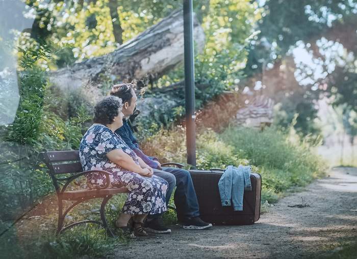 Pokaż zdjęcie: Pan i Pani siedzą na ławce z torbami podróżnymi i patrzą w dal