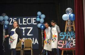 Pokaż zdjęcie: Dwie kobiety oraz mężczyzna stoją na sali. Mają na sobie ręcznie malowane fartuchy. Po prawej stronie na sztaludze ustawiony został napis: „Szansa na sukces”. W tle widoczny jest też napis: „25-lecie WTZ w Rydzynie” udekorowany niebieskimi balonami.