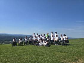 Pokaż zdjęcie: Na trawiastym wzgórzu, na tle gór oraz bezchmurnego nieba znajduje się grupa około dwudziestu osób. Część z tych osób porusza się na wózku.