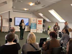 Pokaż zdjęcie: Prelegentka stoi przed publicznością w sali konferencyjnej, gestykulując podczas wystąpienia. Za nią ekran z prezentacją oraz roll-upy Porozumienia Autyzm Polska i Stowarzyszenia Pomocy Osobom Autystycznym. Publiczność słucha uważnie.  