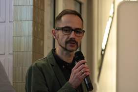 Pokaż zdjęcie: Szczupły mężczyzna w okularach, wąsami i bródką ubrany jest w zieloną marynarkę. Zdjęcie obejmuje górną część ciała, w ręku trzyma mikrofon