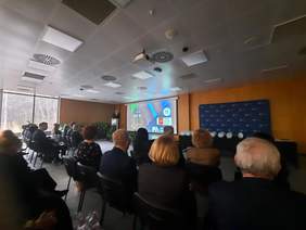 Pokaż zdjęcie: W sali siedzą na krzesłach ludzie. Patrzą na ekran, na którym wyświetlona jest prezentacja. Przy mównicy stoi mężczyzna, który przemawia do mikrofonu. 