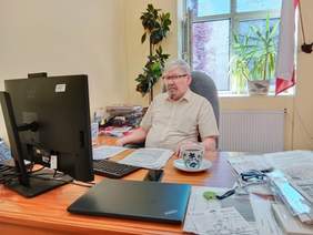 Pokaż zdjęcie: Przy biurku siedzi dyrektor Łódzkiego Oddziału PFRON. Przed nim znajduje się komputer. Na biurku leżą różne dokumenty, filiżanka z herbatą, telefon, klucze. 