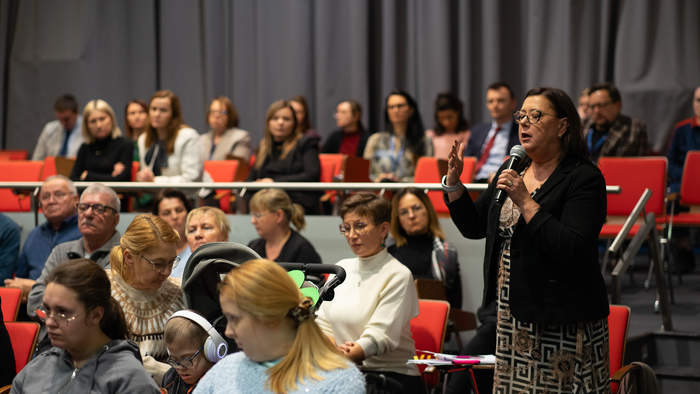 Zdjęcie przedstawia słuchaczy, zgromadzonych na sali konferencyjnej. Po prawej stronie stoi kobieta, która trzyma mikrofon w dłoni i przemawia