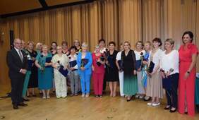 Pokaż zdjęcie: Grupa kobiet w eleganckich strojach pozuje z różami i dyplomami na tle drewnianej ściany, obok stoi mężczyzna w garniturze.