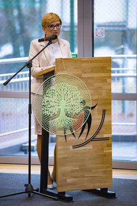 Pokaż zdjęcie: Kobieta w jasnym kostiumie przemawia zza drewnianej mównicy, na której wisi okrągła dekoracja w kształcie stylizowanego, jasnozielonego drzewa.