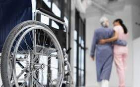 Zdjęcie wózka inwalidzkiego, a w tle dwie osoby - chora i asystent  