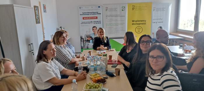 Pokaż zdjęcie: Goście spotkania siedzą przy stolikach, na których jest kawa i poczęstunek