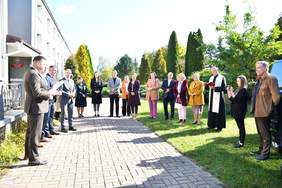 Pokaż zdjęcie: Na zewnątrz przy aucie stoją zaproszeni goście, ksiądz, władze samorządowe, pracownicy i mieszkanki ośrodka.