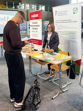 Pokaż zdjęcie: Młody mężczyzna podchodzi do stoiska informacyjnego, przegląda materiały. Kobieta przy stoisku rozmawia z zainteresowanym. Za stoiskiem stoi rollup z logo PFRON.