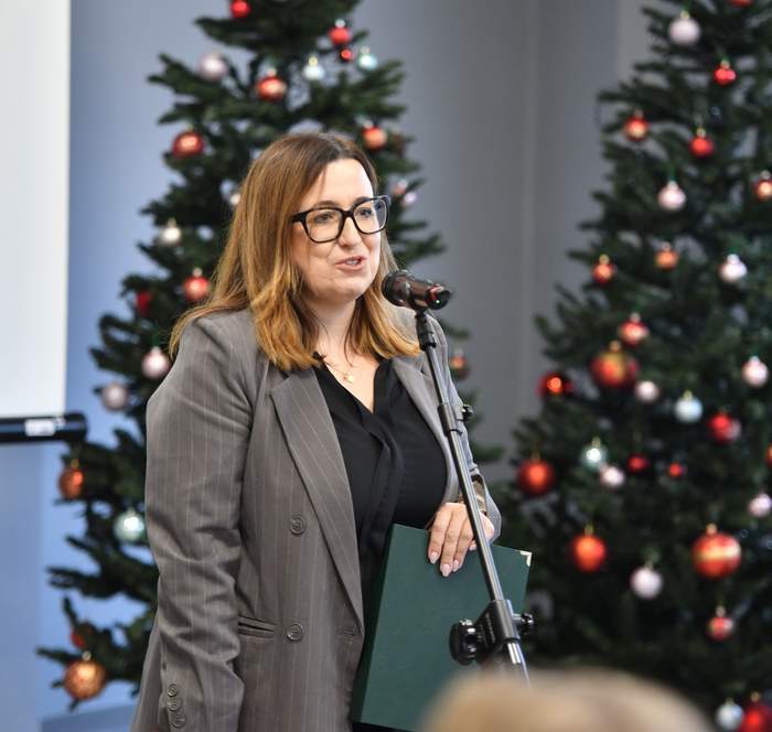 Pokaż zdjęcie: Przy mikrofonie stoi kobieta w okularach, lekko się uśmiecha.