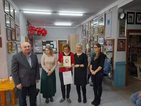 Pokaż zdjęcie: W pracowni artystycznej Warsztatu Terapii Zajęciowej pozuje do pamiątkowego zdjęcia 5 osób. Pośrodku stoi kobieta w czerwonej bluzce, w ręku trzyma dyplom oraz siatkę z nagrodami. Po lewej stronie zdjęcia stoją pracownicy PFRON mężczyzna i kobieta, po prawej dwie kobiety pracujące w WTZ w Chełmie.