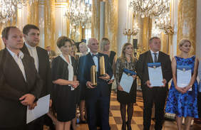 Pokaż zdjęcie: Grupa osób ustawionych w rzędzie trzyma w rękach dyplomy i statuetki