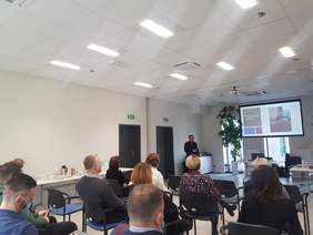 Sala konferencyjna. Na krzesłach siedzą przedstawiciele podmiotów publicznych. Przed uczestnikami stoi pan Łukasz Żmuda - Dyrektor Oddziału Opolskiego PFRON. Na ekranie na ścianie wyświetlana jest prezentacja dot. składania skarg na brak dostępn