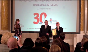 Pokaż zdjęcie: Na scenie Pan Paweł Wdówik – Pełnomocnik Rządu ds. Osób Niepełnosprawnych odbiera wyróżnienie IDOLA 30-LECIA od Pana Marka Kalbarczyka. W tle telebim z logotypem fundacji. W dole zdjęcia, siedząca publiczność