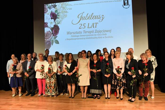 Pokaż zdjęcie: Na scenie do pamiątkowego zdjęcia ustawili się wszyscy pracownicy Warsztatu Terapii Zajęciowej , osoby te trzymają w ręku po jednej czerwonej róży. Nad ich głowami jest wyświetlana plansza z napisem Jubileusz 25-lecie WTZ