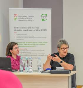 Pokaż zdjęcie: Dwie kobiety siedzą przy stole podczas spotkania informacyjnego PFRON. Na drugim planie widoczny jest baner Centrum informacyjno-doradczego dla osób z niepełnosprawnością (CIDON).