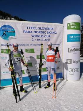 Dwóch narciarzy stoi na tle baneru mistrzostw, uśmiechają się, tło słoneczne