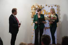 Pokaż zdjęcie: Od lewej mężczyzna następnie kobieta w zielonej marynarce trzyma w ręku grawerton obok stoi wysoka blondynka trzymająca w ręku bukiet kwiatów.