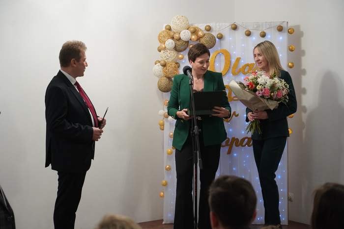 Pokaż zdjęcie: Od lewej mężczyzna następnie kobieta w zielonej marynarce trzyma w ręku grawerton obok stoi wysoka blondynka trzymająca w ręku bukiet kwiatów.