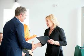 Pokaż zdjęcie: Na zdjęciu kobieta , blondynka w czarnej sukience składa życzenia  mężczyźnie w garniturze