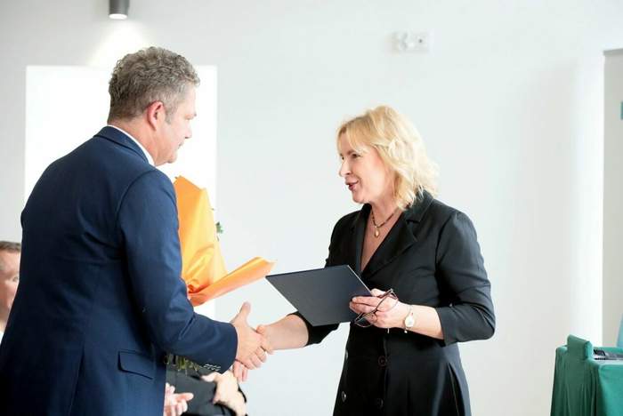 Pokaż zdjęcie: Na zdjęciu kobieta , blondynka w czarnej sukience składa życzenia  mężczyźnie w garniturze