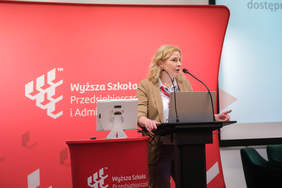 Pokaż zdjęcie: Zdjęcie mównicy przy której stoi kobieta, blondynka w okularach w beżowym garniturze.