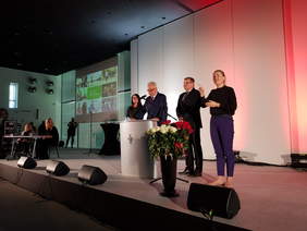 Pokaż zdjęcie: Widok na scenę, przy mikrofonie przemawia mężczyzna w garniturze i krawacie, obok stoi kobieta w czarnej bluzce i granatowych spodniach, tłumacz języka migowego. Za przemawiającym mężczyzną stoi kobieta w okularach i mężczyzna w garniturze. Na 