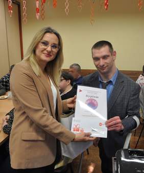 Pokaż zdjęcie: Kobieta wręcza dyplom mężczyźnie w sali konferencyjnej. Na dokumencie widoczna jest okrągła, kolorowa grafika. W tle siedzą uczestnicy spotkania, a z sufitu zwisają dekoracyjne, czerwone i różowe serca.