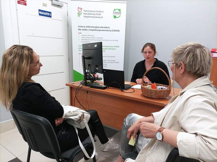 Dwie kobiety siedzą na krzesłach - tyłem do obiektywu - uczestniczki konsultacji. Przodem do nich, przy biurku z komputerem siedzi pracownik PFRON. W tle widać banery.