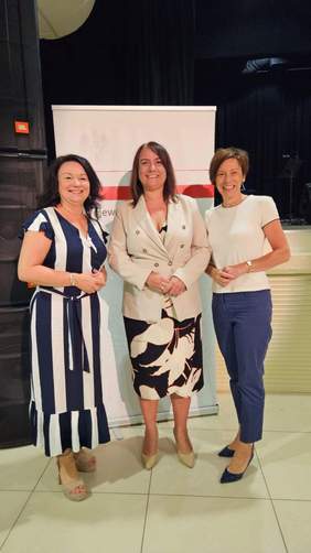 Pokaż zdjęcie: Trzy uśmiechnięte kobiety stoją w sali ze ścianami w kolorze czarnym. Na posadzce ułożone są kremowe płytki. W tle znajduje się baner wojewody wielkopolskiego.