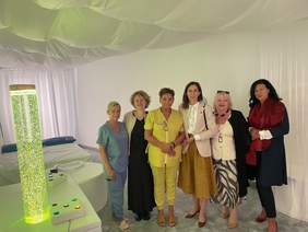 Pokaż zdjęcie: Grupa sześciu kobiet stoi razem w jasnym, nowoczesnym pomieszczeniu wyposażonym w sprzęt rehabilitacyjny i relaksacyjny.