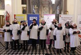 Pokaż zdjęcie: na zdjęciu  miejsce w kosciele po środku stoją osoby ubrane na biało w białe stroje  stoją i śpiewają dla publiczności 