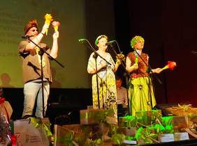 Pokaż zdjęcie: Na scenie trzech wykonawców - dwie kobiety, jeden mężczyzna. Wszyscy są kolorowo ubrani i śpiewają.