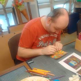 Pokaż zdjęcie: Fragment pomieszczenia w którym przy stole siedzi mężczyzna w czerwonej koszulce i maluje kartkę świąteczną.