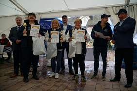 Pokaż zdjęcie: Pod zadaszeniem w rzędzie stoi 7 osób 3 kobiety ubrane na sportowo trzymają w rękach dyplomy, statuetki i reklamówki z upominkami