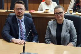 Pokaż zdjęcie: Portretowe ujęcie dwóch osób siedzących obok siebie za drewnianym stołem podczas konferencji: mężczyzny w granatowym garniturze i fioletowo-białym krawacie oraz kobiety w szarym, tweedowym żakiecie. Oboje mają okulary i patrzą prosto w obiektyw. Przed nimi znajduje się mikrofon.